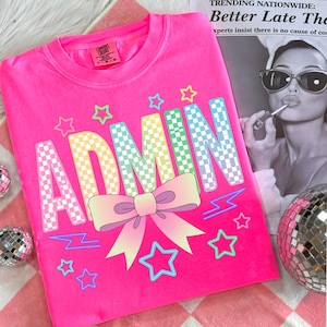 Può includere: Una felpa rosa acceso con la scritta "ADMIN" in lettere a scacchi colorate. Un fiocco pastello e grafiche a stella decorano il davanti. La maglietta è su una superficie a scacchi rosa e bianca.