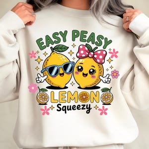 Puede incluir: Sudadera color crema con un diseño de limón de dibujos animados. Un limón lleva gafas de sol, el otro un lazo rosa con lunares. El texto "EASY PEASY LEMON SQUEEZY" está impreso encima y debajo de los limones, con detalles florales y estrellas.