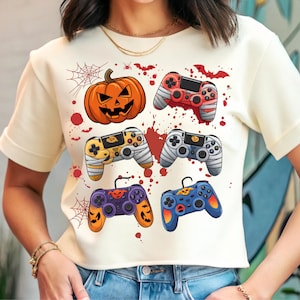 Op de afbeelding: Crèmekleurig T-shirt met een Halloween-thema. De afbeelding bevat een pompoen, gamecontrollers verpakt in verbanden en controllers met pompoen- en spinnenwebontwerpen. Rode bloedspetters en spinnenwebben maken het griezelige thema compleet.