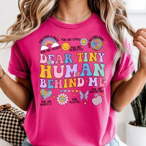May include: A vibrant pink t-shirt with the words "DEAR TINY HUMAN BEHIND ME" in a retro font. The design includes a rainbow, hearts, flowers, and a butterfly, alongside positive affirmations. A cheerful and colourful design.