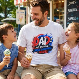 May include: A white short-sleeved t-shirt featuring a graphic print with the text DAD: THE ORIGINAL LARGE LANGUAGE MODEL in red, white, and blue colours with a paint splatter effect and a comic book style dot pattern background.
