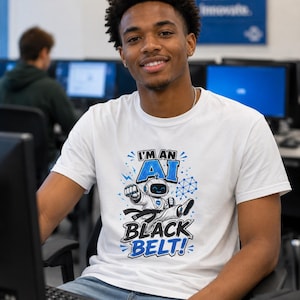 May include: White t-shirt with the text "I'M AN AI BLACK BELT!" in blue and black. The graphic features a cartoon robot in a karate pose. The person is sitting in front of a computer.