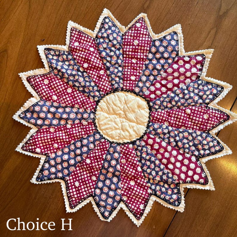 May include: A handmade, quilted sunflower-shaped trivet. The petals feature a mix of patterned fabrics in burgundy and navy blue, with a cream-colored center. The trivet is edged with a white, scalloped trim and black stitching. The text "Choice H" is visible.