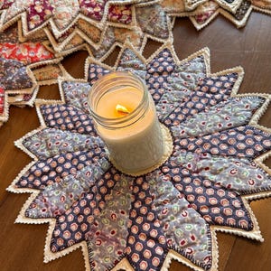 May include: A handmade, star-shaped quilted table mat with a candle in a glass jar. The mat features a patchwork design with various floral and geometric patterns in shades of blue, purple, and cream. The candle is lit, casting a warm glow.