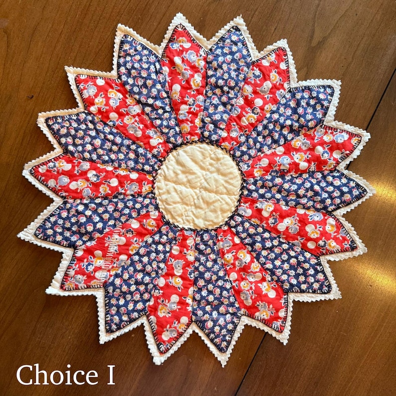 May include: A handmade, flower-shaped quilted trivet. The trivet features alternating red and navy blue fabric petals with a cream-colored center. The petals are edged with white lace and the text "Choice I" is visible.
