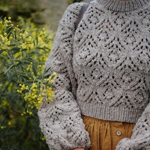 May include: A light grey knitted jumper featuring an intricate openwork design. The jumper has long, puffed sleeves and a ribbed hem. The person is wearing a mustard-coloured skirt. Yellow flowers are in the background.