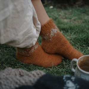Puede incluir: Primer plano de los pies de una persona con calcetines de punto color óxido con un diseño blanco. Los calcetines están sobre hierba verde. Una taza blanca y una manta están en primer plano.