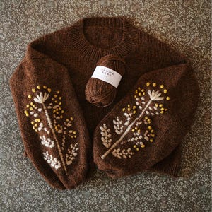 May include: A brown knitted jumper with embroidered floral patterns on the sleeves. The embroidery includes white and yellow details. A ball of brown yarn with a white label is next to the jumper. The label reads "RAUMA GARN".