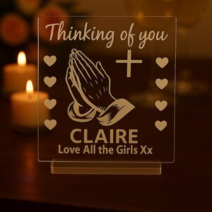 Könnte beinhalten: Transparente Acrylplatte mit den eingravierten Worten "Thinking of you" und "CLAIRE". Das Design umfasst betende Hände, ein Kreuz und Herzsymbole. Der Text "Love All the Girls Xx" ist ebenfalls vorhanden.