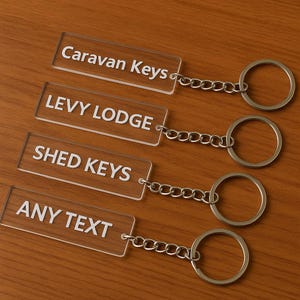 May include: Four clear acrylic keychains with silver chains and key rings. Each keychain has a rectangular shape with white text: "Caravan Keys", "LEVY LODGE", "SHED KEYS", and "ANY TEXT". The keychains are displayed on a wooden surface.