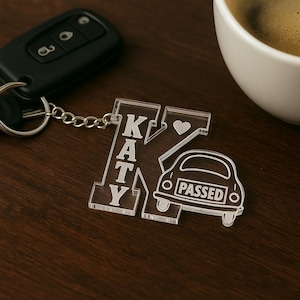 Pode incluir: Porta-chaves em acrílico transparente com o nome KATY e um carro com a palavra PASSED. O porta-chaves está preso a um porta-chaves de carro com uma corrente prateada. Uma chávena de café branca está no fundo.