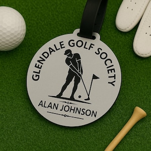 May include: A circular light grey luggage tag with a black strap. The tag features a black graphic of a golfer, the text GLENDALE GOLF SOCIETY, and the name ALAN JOHNSON printed in black. The design includes a golf flag and a small ball.