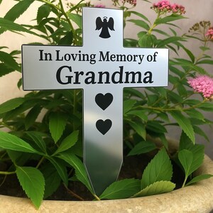 Puede incluir: Una estaca conmemorativa de metal plateado en forma de cruz con las palabras "In Loving Memory of Grandma" en negro. La estaca presenta una silueta de ángel negro y dos corazones negros. La cruz está colocada en una maceta con plantas verdes y flores rosas.