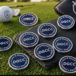 May include: A collection of round, blue and silver metal golf ball markers featuring the text Luke & Niamh's Captain's Day 2026 and DGCC. The markers are displayed on a dark slate surface alongside a white golf ball with a matching logo.