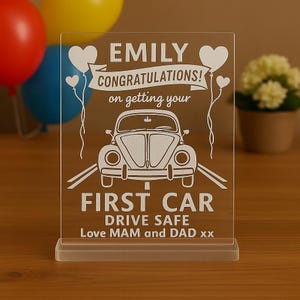 May include: A clear acrylic plaque with the text "EMILY CONGRATULATIONS! on getting your FIRST CAR DRIVE SAFE Love MAM and DAD xx" and a car graphic. The plaque has a rectangular shape and a small base. Balloons are in the background.