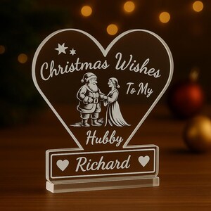 Peut inclure: Décoration de Noël en acrylique transparent en forme de cœur. L'ornement présente une image gravée du Père Noël et d'une femme, avec le texte "Christmas Wishes To My Hubby Richard". Deux cœurs encadrent le nom.