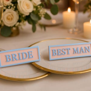 May include: A wedding table setting featuring two plates with gold rims. Each plate has a sign: one reads "BRIDE", the other "BEST MAN". The signs are light blue on a pink background. Champagne glasses, flowers, and candles are visible in the background.