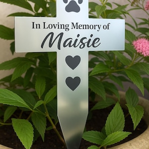 May include: A silver metal pet memorial stake in a cross shape, with the words "In Loving Memory of Maisie". The stake features a paw print and two heart symbols. The memorial is placed in a pot with green plants and pink flowers.