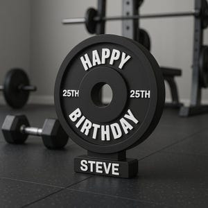 Op de afbeelding: Een zwarte halterplaat met de woorden "HAPPY BIRTHDAY STEVE" in witte letters. Het nummer "25th" staat aan weerszijden van het middelste gat. De halterplaat staat op een zwart oppervlak met fitnessapparatuur op de achtergrond.