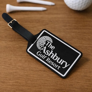 May include: Black luggage tag with the text "The Ashbury Golf Resort" in white. The tag has a black strap with a gold-coloured buckle. Two white golf tees and a golf ball are also visible.