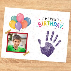 Op de afbeelding: Een rechthoekige verjaardagskaart met een tros gekleurde ballonnen, een foto van een kind in een groen shirt in een rode lijst en een paarse handafdruk. De tekst "happy BIRTHDAY" staat in gekleurde letters met kleine sterretjes en hartjes.