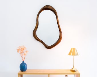 Puede incluir: Una mesa consola de madera con un espejo único de forma orgánica colgado encima. La mesa tiene dos estantes y una lámpara dorada con una pantalla blanca se encuentra en el estante superior.