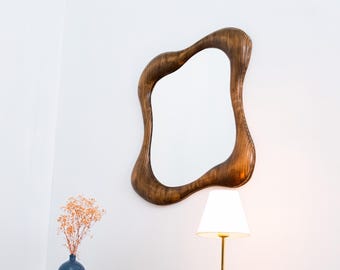 Puede incluir: Un espejo de madera con forma orgánica y acabado marrón cuelga de una pared blanca. El marco del espejo tiene una apariencia texturizada. Debajo, hay un pequeño jarrón azul con flores secas y una lámpara de latón con pantalla blanca.