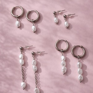 Può includere: Una collezione di orecchini argentati con dettagli di perle bianche, presentati su uno sfondo rosa. Gli orecchini includono modelli a cerchio e pendenti con diverse quantità di perle. Le perle hanno una finitura lucida.