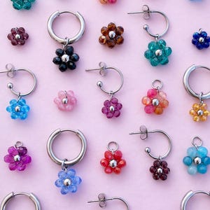 Op de afbeelding: Een verzameling kleurrijke bloemvormige oorbellen van kralen in verschillende tinten, waaronder blauw, groen, rood en paars. Sommige oorbellen zijn creolen, andere zijn oorstekers. De oorbellen worden gepresenteerd op een lichtroze achtergrond.