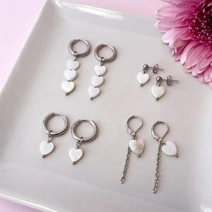 Op de afbeelding: Een collectie hartvormige oorbellen in verschillende stijlen. De oorbellen hebben witte hartvormige hangers en zilverkleurige metalen ringen en pinnen. Sommige oorbellen hebben meerdere harten, andere hebben één hartvormige hanger. Een roze bloem is op de achtergrond.