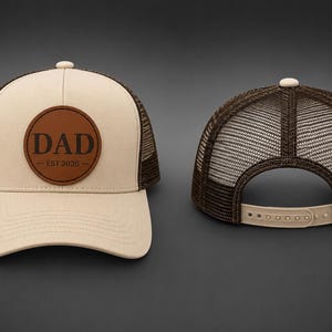 Peut inclure: Une casquette trucker beige et marron avec un écusson circulaire en cuir marron sur le panneau avant portant le texte DAD EST 2026. La casquette possède un dos en filet, une visière incurvée et une fermeture snapback réglable à l'arrière.