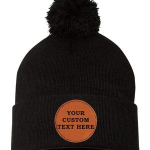 Peut inclure: Un bonnet noir en tricot avec un patch en cuir marron qui dit "YOUR CUSTOM TEXT HERE" et un pompon noir sur le dessus.