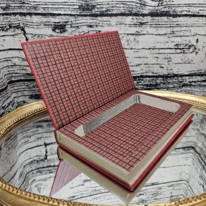May include: A hollowed-out book with a red cover featuring a black grid pattern. The interior pages are cut out to create a rectangular storage compartment with silver-toned edges, revealing the grid design on the bottom of the cavity.