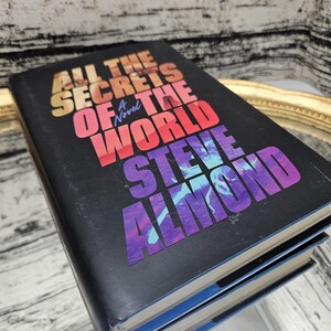 以下が含まれることがあります： スティーブ・アーモンド著「All the Secrets of the World: A Novel」というタイトルのハードカバー本。本の表紙は黒色で、オレンジ、赤、紫色のカラフルな文字が書かれています。本は反射する表面に置かれています。