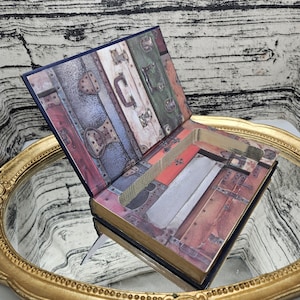 Pode incluir: Uma caixa decorativa em forma de livro com um design de mala vintage na capa e no interior. A caixa está aberta, revelando um espaço oco. A capa apresenta uma colagem de painéis de malas antigas em várias cores, incluindo vermelho, verde e castanho.