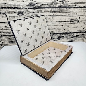 May include: A black book-shaped box with a skull and crossbones pattern on the cover and inside. The box is open, revealing the interior lining.