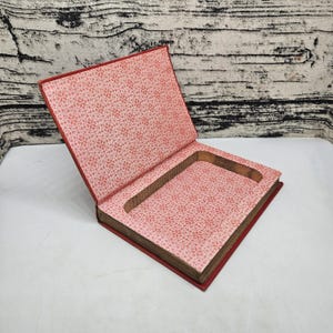 May include: Open red book with a carved-out interior, revealing a hidden compartment. The inside covers feature a pink floral pattern. The book's pages are visible, and the edges are stained.