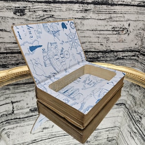 May include: A hollowed-out book with a tan exterior and a white interior lining featuring blue nautical illustrations, including a ship's wheel, an anchor, and the text "Sailor spirit" and "Explorer" printed in a script font.