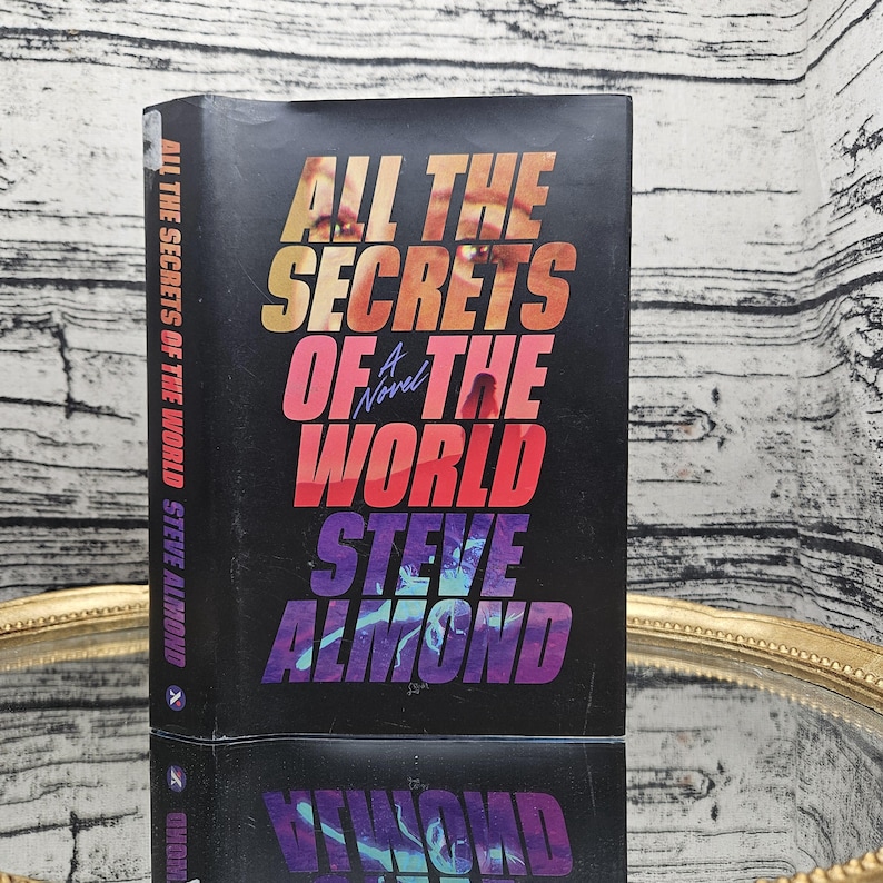 以下が含まれることがあります： スティーブ・アーモンド著「All The Secrets Of The World」というタイトルのハードカバー本。本の表紙は黒色で、タイトルが大きくカラフルな文字で書かれています。本は反射する表面に表示されています。