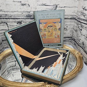 Op de afbeelding: Een open, vintage boek met een uitgehold interieur, met een illustratie van een stadsgezicht. De kaft van het boek luidt "A Picturesque Tale of Progress". Het boek is groen met gouden accenten, en het interieur is zwart met oranje en blauwe details.