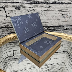 May include: An open, book-shaped box with a blue snowflake-patterned interior. The box has a hollowed-out space and aged edges, giving it a vintage appearance. The box is resting on a reflective surface.