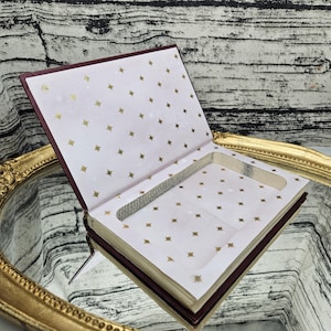 May include: An open book-shaped box with a burgundy cover and gold-edged pages. The interior is lined with white paper adorned with gold star patterns. A rectangular cutout is visible in the center, suggesting a storage compartment.