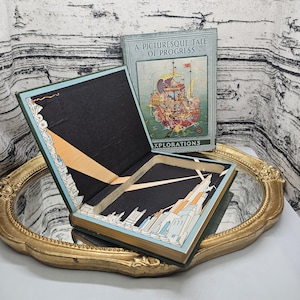Op de afbeelding: Een open, vintage boek met een uitgehold interieur, dat een verborgen compartiment onthult. De omslag van het boek bevat de titel "A Picturesque Tale of Progress" en een illustratie van een schip. Het interieur is versierd met een stadsgezicht.