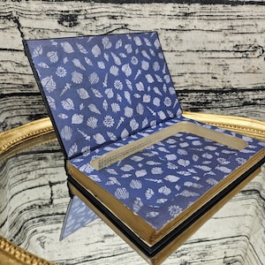 May include: A book with a blue seashell pattern on the inside covers, and gold-edged pages. The book is open, revealing a rectangular space carved out of the pages. The exterior of the book is black.
