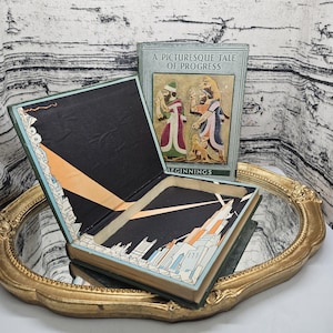 Op de afbeelding: Een open vintage boek met een zwart interieur en een illustratie van een stadsgezicht. De kaft van het boek luidt "A Picturesque Tale of Progress" met een illustratie van figuren. Het boek ligt op een goudkleurige spiegel.