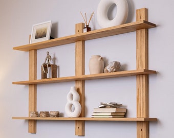 May include: A wooden wall shelf with three shelves. The shelves are filled with decorative items, including a vase, a sculpture, and books. The shelf is made of natural wood and has a simple, modern design.