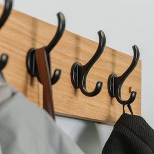May include: A wooden wall-mounted coat rack with five black metal hooks. A black face mask and a brown leather strap are hanging on the hooks.