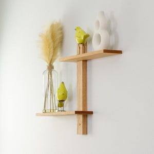 May include: A two-tiered wooden wall shelf with a vertical support. The top shelf holds a white vase and a yellow bird figurine. The bottom shelf displays a clear glass bottle with dried pampas grass and another yellow bird figurine.