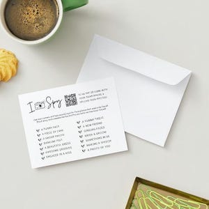 May include: A white card with a black camera icon and the text "I Spy" printed on it. The card has a list of things to find, such as "a funny face", "a piece of cake", "a group photo", "dancing feet", "a beautiful dress", "awesome grooves", "engaged in a kiss", "a yummy treat", "a new friend", "singing faces", "rice & gravy", "something blue", "making a speech", and "a photo of you". There is a QR code on the card.