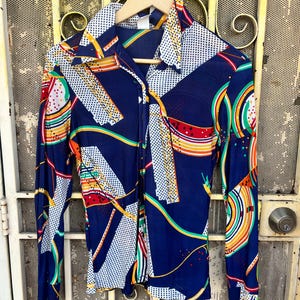 Puede incluir: Una camisa vintage de manga larga con un vibrante estampado geométrico. La camisa presenta una base azul marino con lunares blancos y acentos coloridos en rojo, amarillo, verde y naranja. Tiene cuello y se abotona por delante.